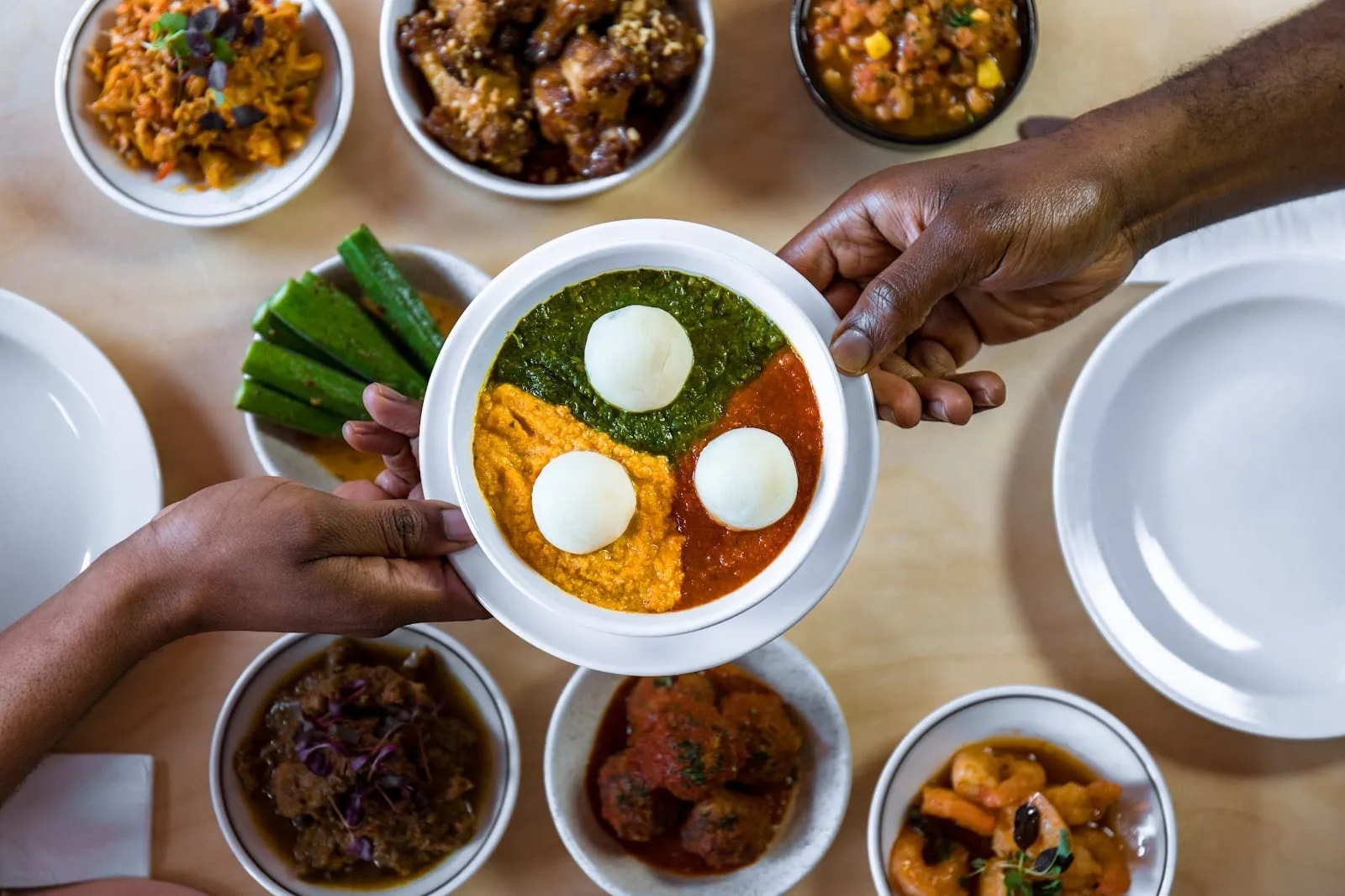 An overhead spread of Nigerian small plates: a central bowl divided between a green stew, a yellow-orange one and a red one, each with a smooth white ball of pounded yam on it, held between two pairs of hands, with okra spears, jollof rice, peanut-crusted wings, meatballs in tomato and prawns in bowls around it