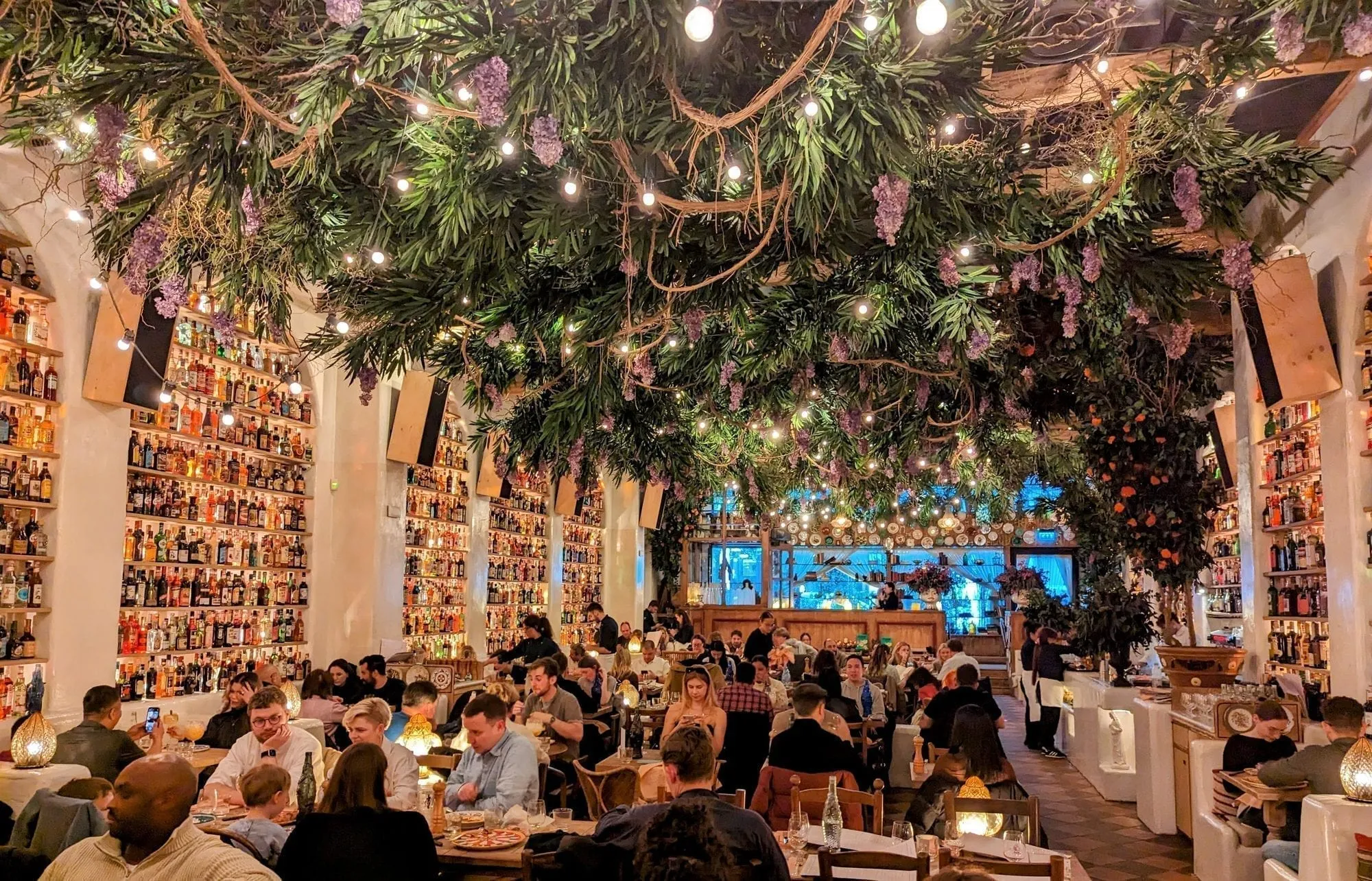 The dining room at Circolo Popolare, walls stacked floor to ceiling with bottles under a ceiling of hanging greenery, wisteria and fairy lights
