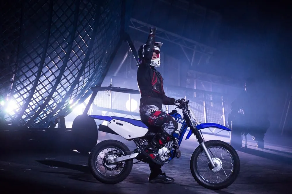 A rider in full leathers and helmet on a motorcycle in front of the steel mesh sphere of the Globe of Death, one arm raised, backlit through stage haze