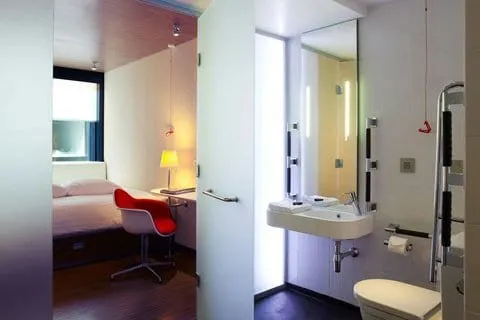 The bathroom of an accessible room at citizenM London Bankside, with grab rails by the toilet, a wall-hung basin and a view through the door to the bed and a red chair