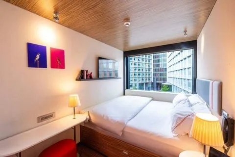 A room at citizenM London Bankside with the bed filling the space from wall to wall under a floor-to-ceiling window, a wood-slatted ceiling and two bird prints