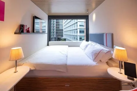 A compact citizenM room with a city-view window, the bed filling almost the full width, and a floating shelf beside it
