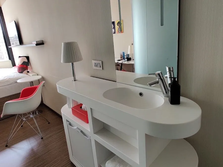 The white washbasin unit standing in the bedroom at citizenM Tower of London, with a lamp and shelves, and the frosted shower pod reflected in the mirror