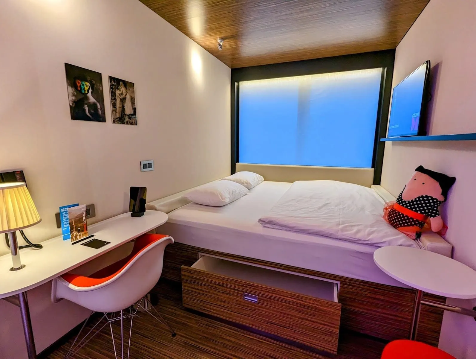 A citizenM Tower of London room with the bed filling the width of the room under a wide window lit blue by the mood lighting, a drawer under the bed, a white desk with a red Eames chair and a TV on a shelf