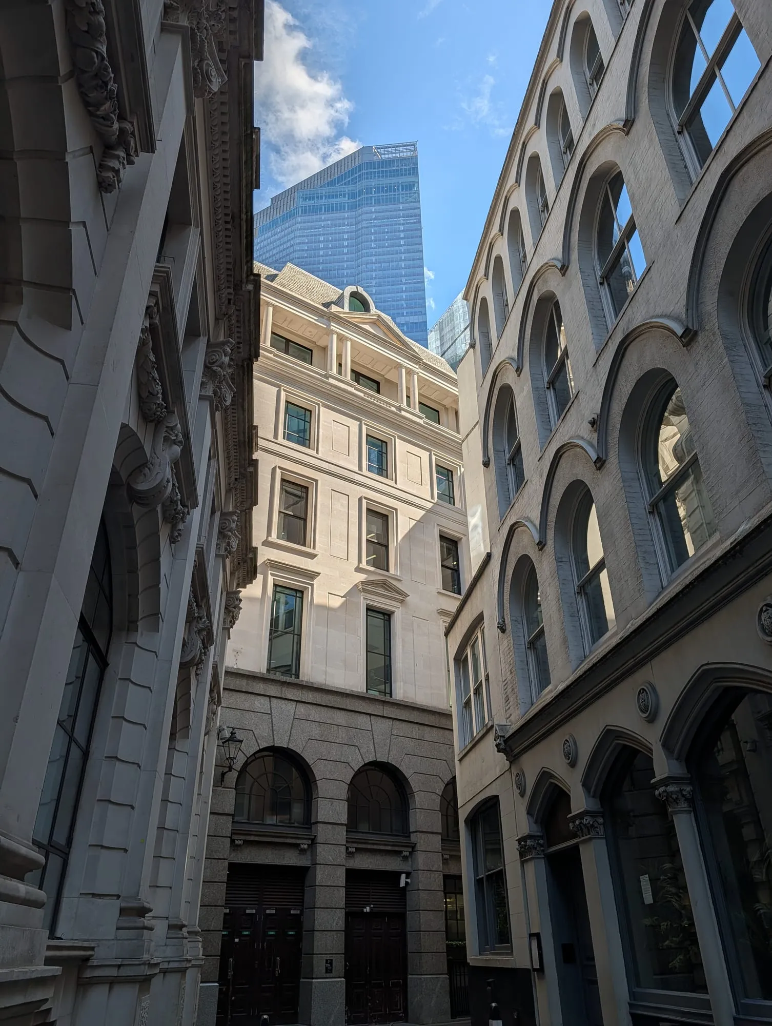 Looking up a narrow City alley, Victorian arched windows on one side and classical stonework on the other, with a glass tower filling the gap of sky above