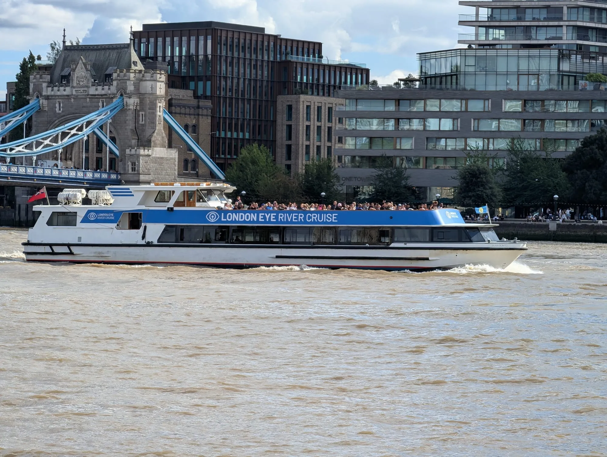 A London Eye River Cruise boat on the Thames with its top deck full, passing the buildings near Tower Bridge