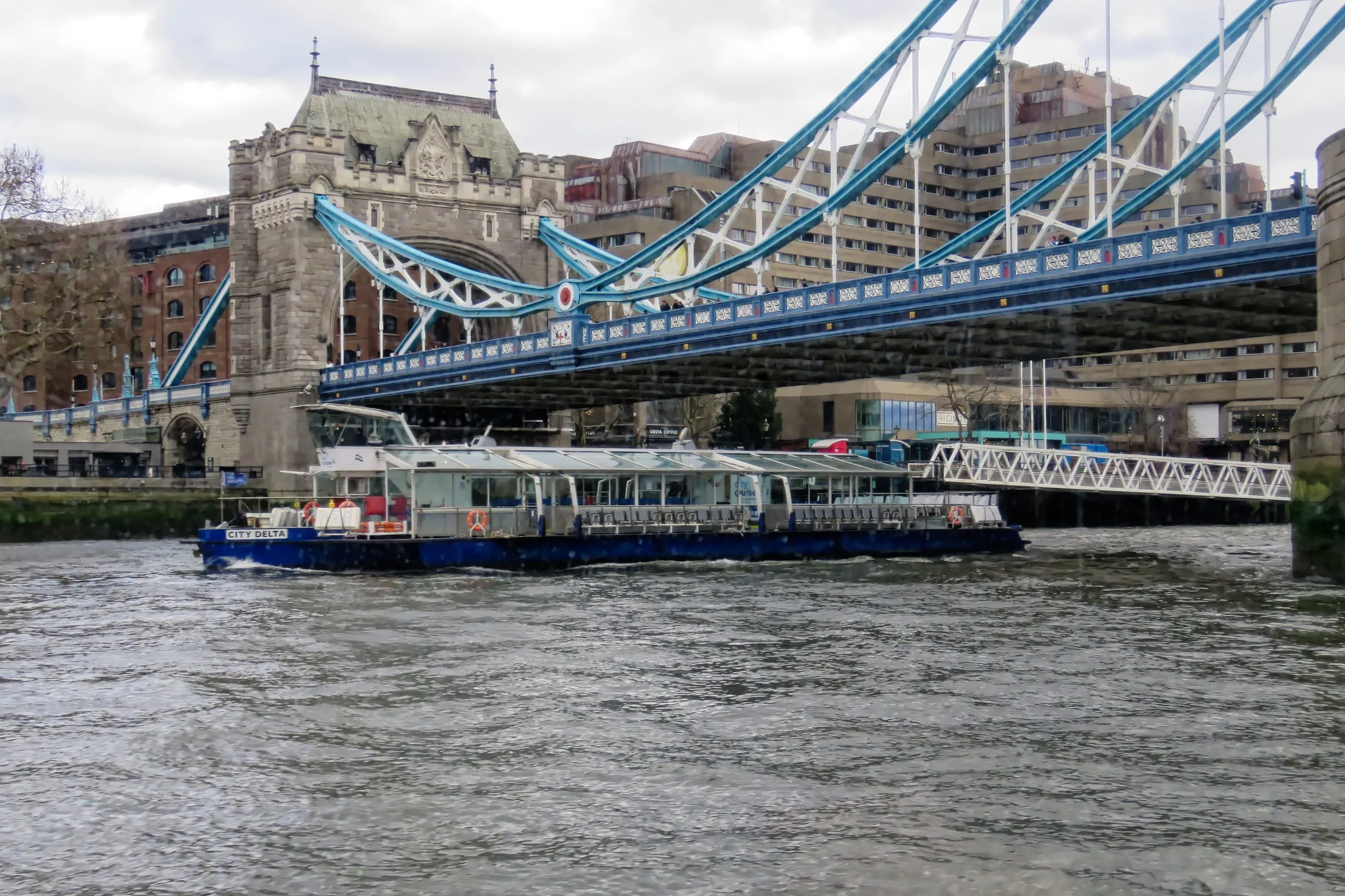The City Cruises sightseeing boat City Delta, with a glass-roofed saloon and an open rear deck, passing beneath Tower Bridge on the Thames