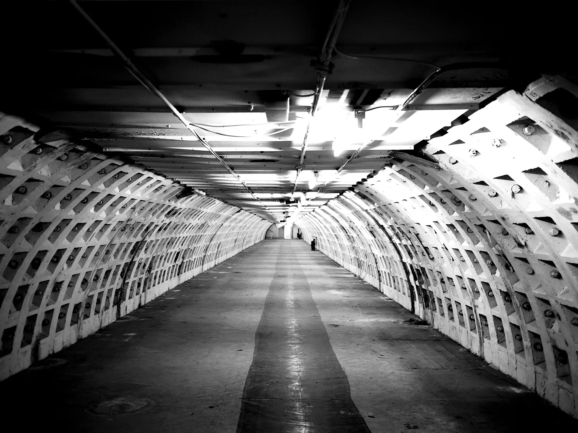 A long arched tunnel in black and white, its walls ribbed with bolted segments and a pale strip down the middle of the floor