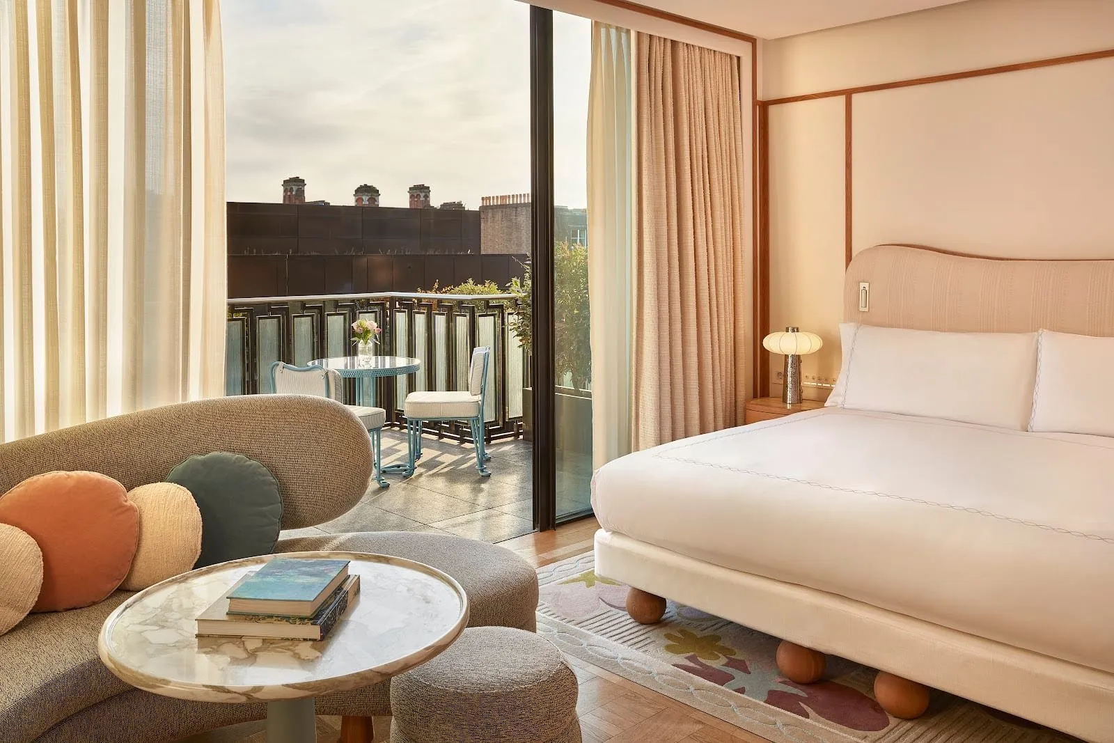 A contemporary Claridge's room with a curved sofa and round cushions, a scalloped cream headboard, a bed on wooden ball feet, and a glass door open onto a balcony with a blue metal table and two chairs above the rooftops