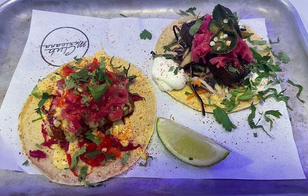 Two loaded tacos on a metal tray at Club Mexicana, piled with pickled red onion, jalapenos, coriander, crema and chilli sauce