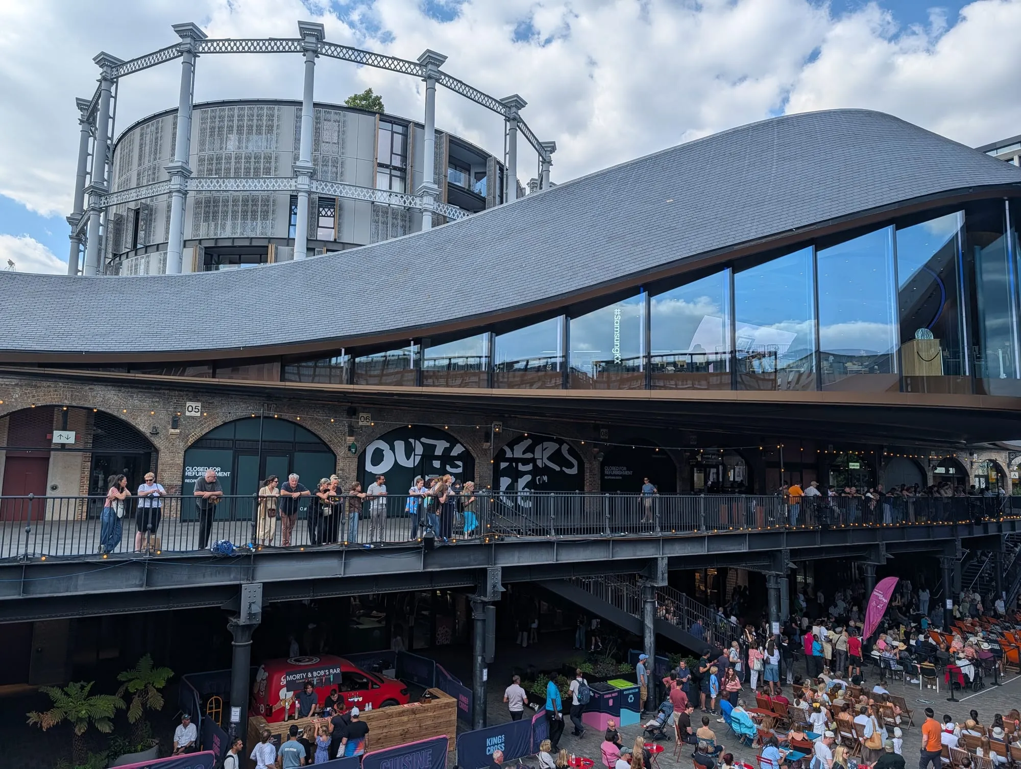 The curving zinc kissing-roof of Coal Drops Yard, where two Victorian warehouse roofs rise to meet each other, above crowds on the upper walkway