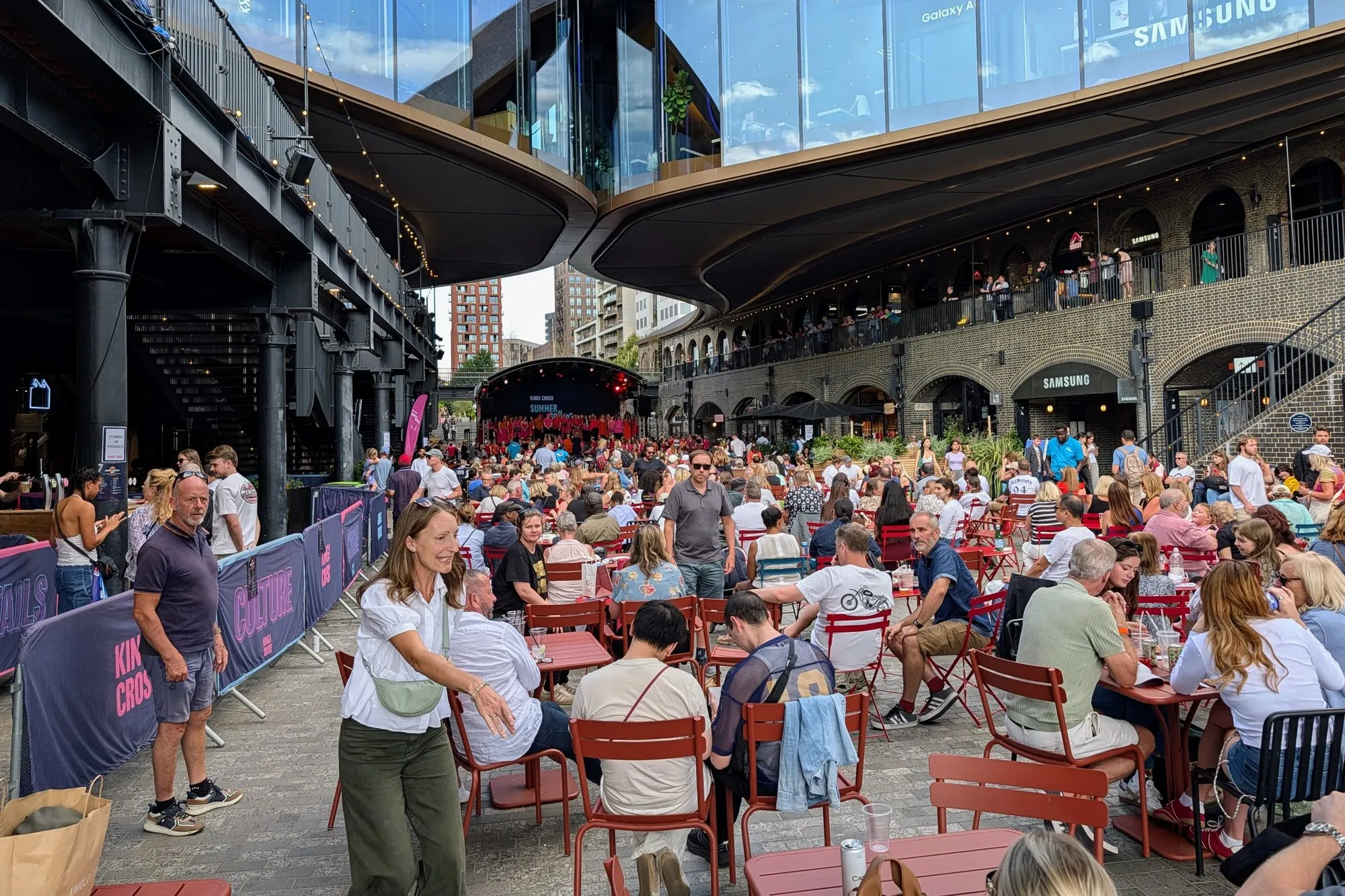 Crowds eating at red bistro tables under the curved roofs of Coal Drops Yard, with a summer stage at the far end and people on the brick arch balconies above
