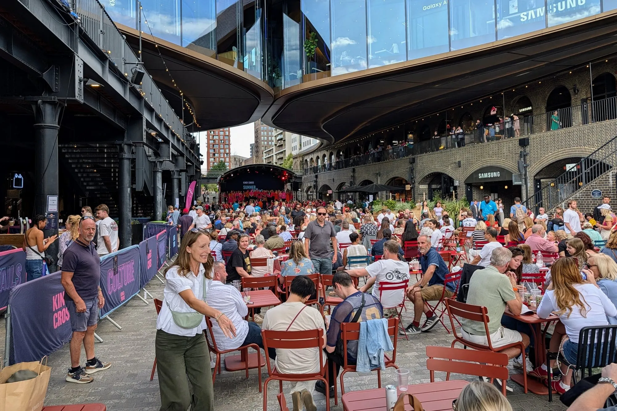 Crowds eating at red bistro tables under the curved roofs of Coal Drops Yard, with a summer stage at the far end and people on the brick arch balconies above