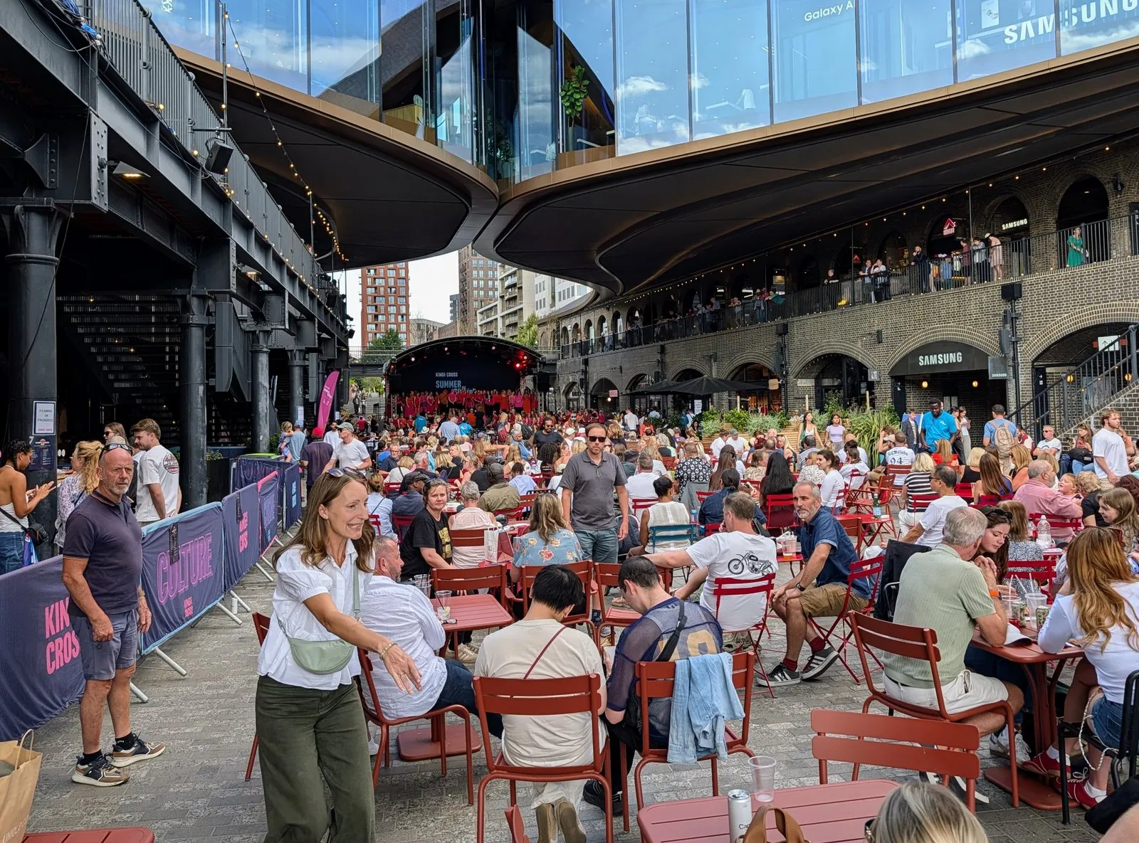 Red bistro tables filled with people eating outside at Coal Drops Yard, under the curved warehouse roofs with brick arch balconies above