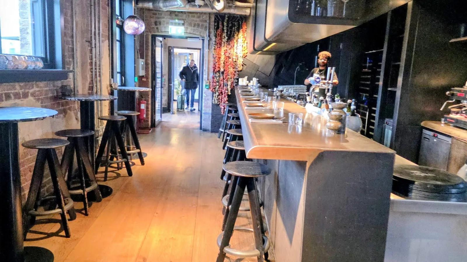 A long bar with stools and an open kitchen inside a brick-walled room