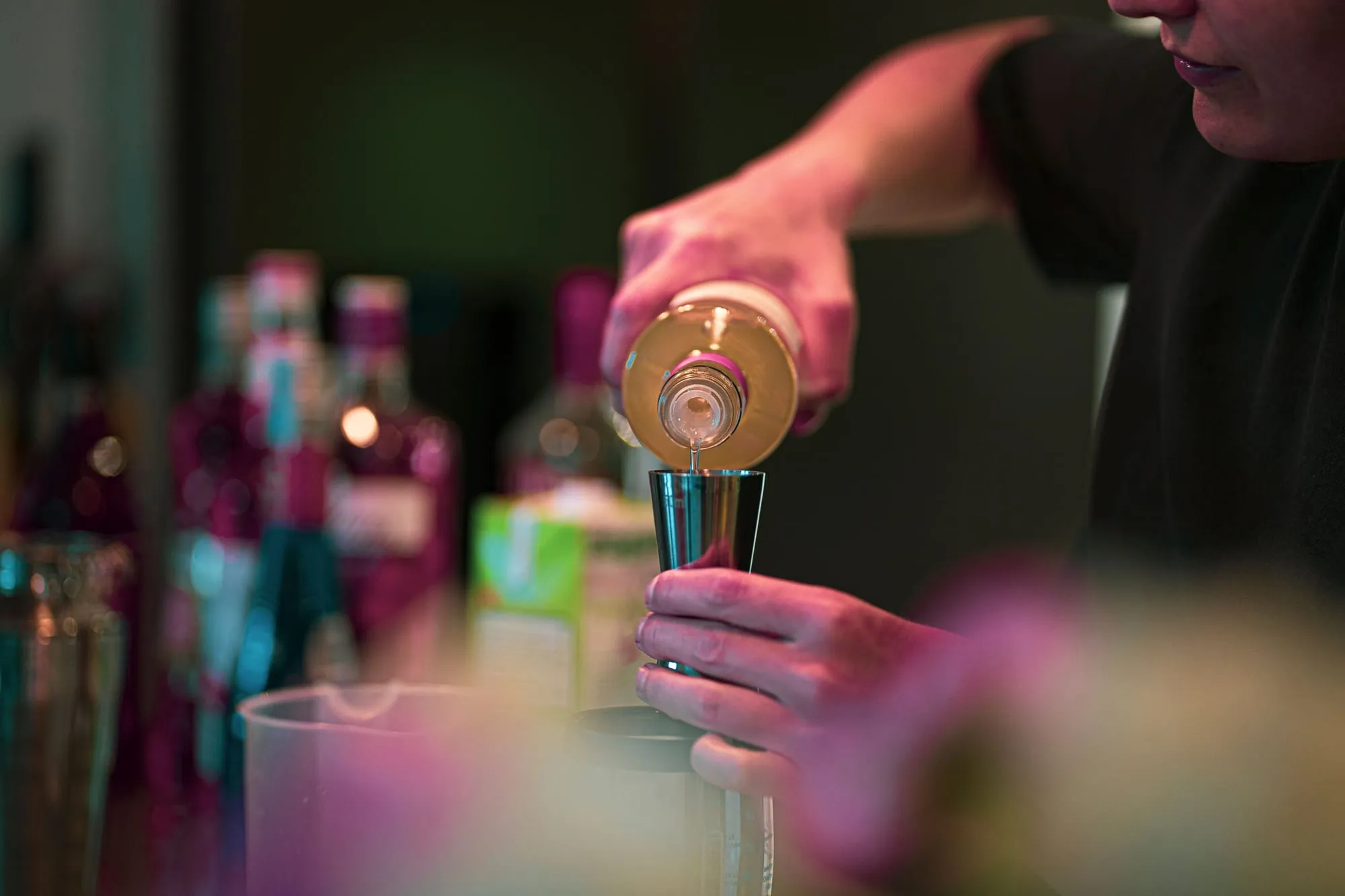 A bartender pouring a measure into a cocktail shaker behind a bar lit pink and teal