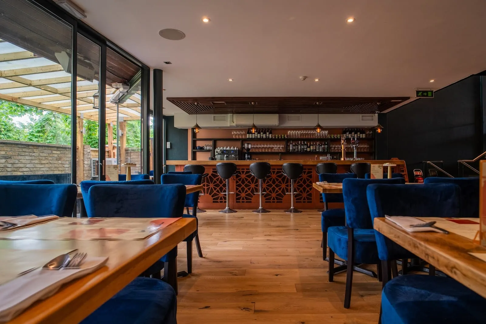 A dining room of blue velvet chairs at light wooden tables, a bar with black stools and a terracotta fretwork front, bottles and glassware on the back shelves, and glass doors folded back onto a covered terrace