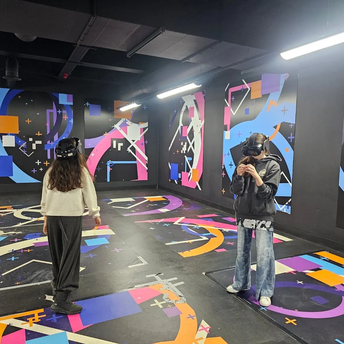 Two people wearing VR headsets standing in a room with a colourful abstract floor and wall graphics, one gesturing with her hands