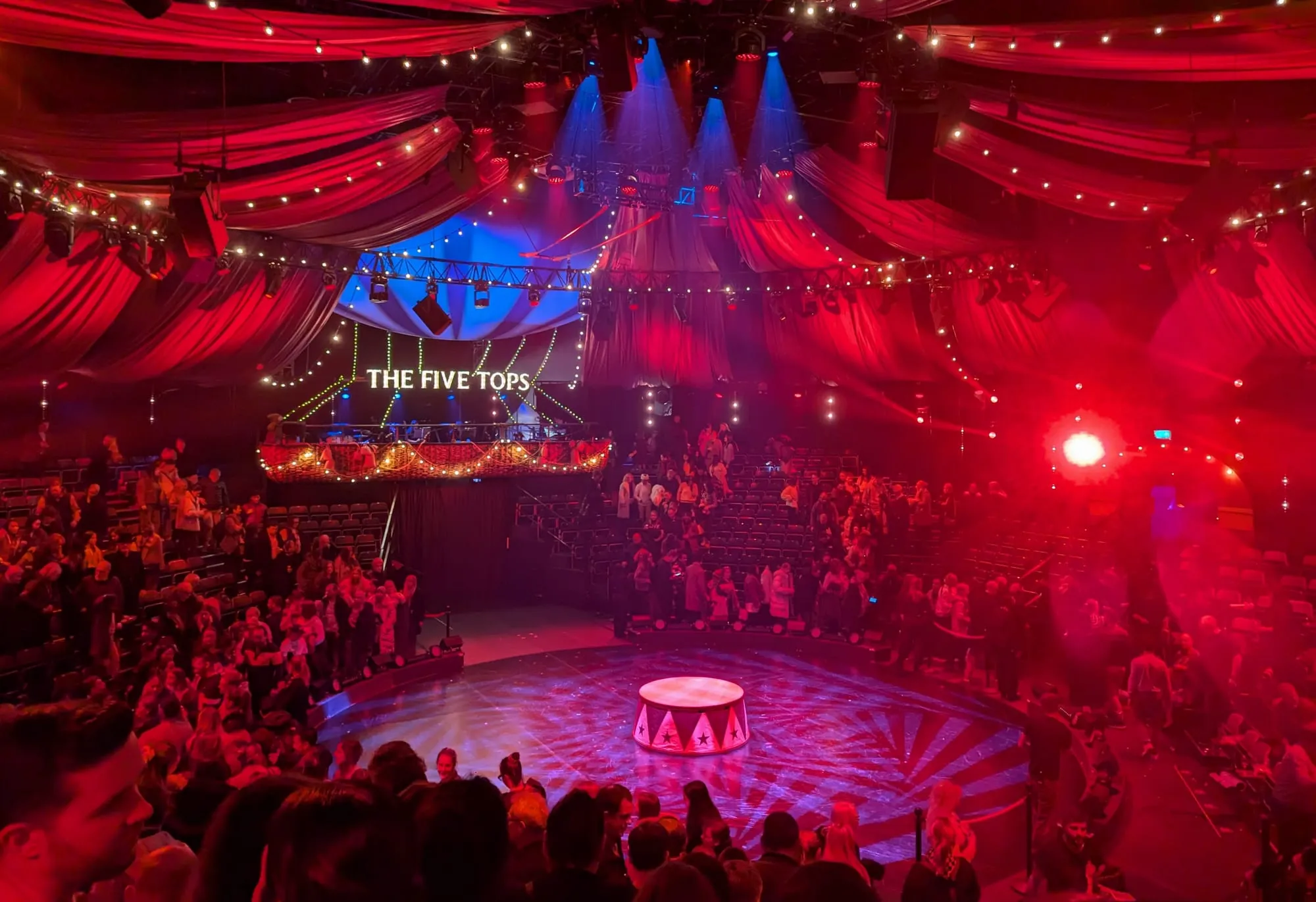 Inside the Big Top at COME ALIVE, the tent draped in red fabric and strung with festoon lights above a circular circus ring, with tiered seating and a sign reading The Five Tops