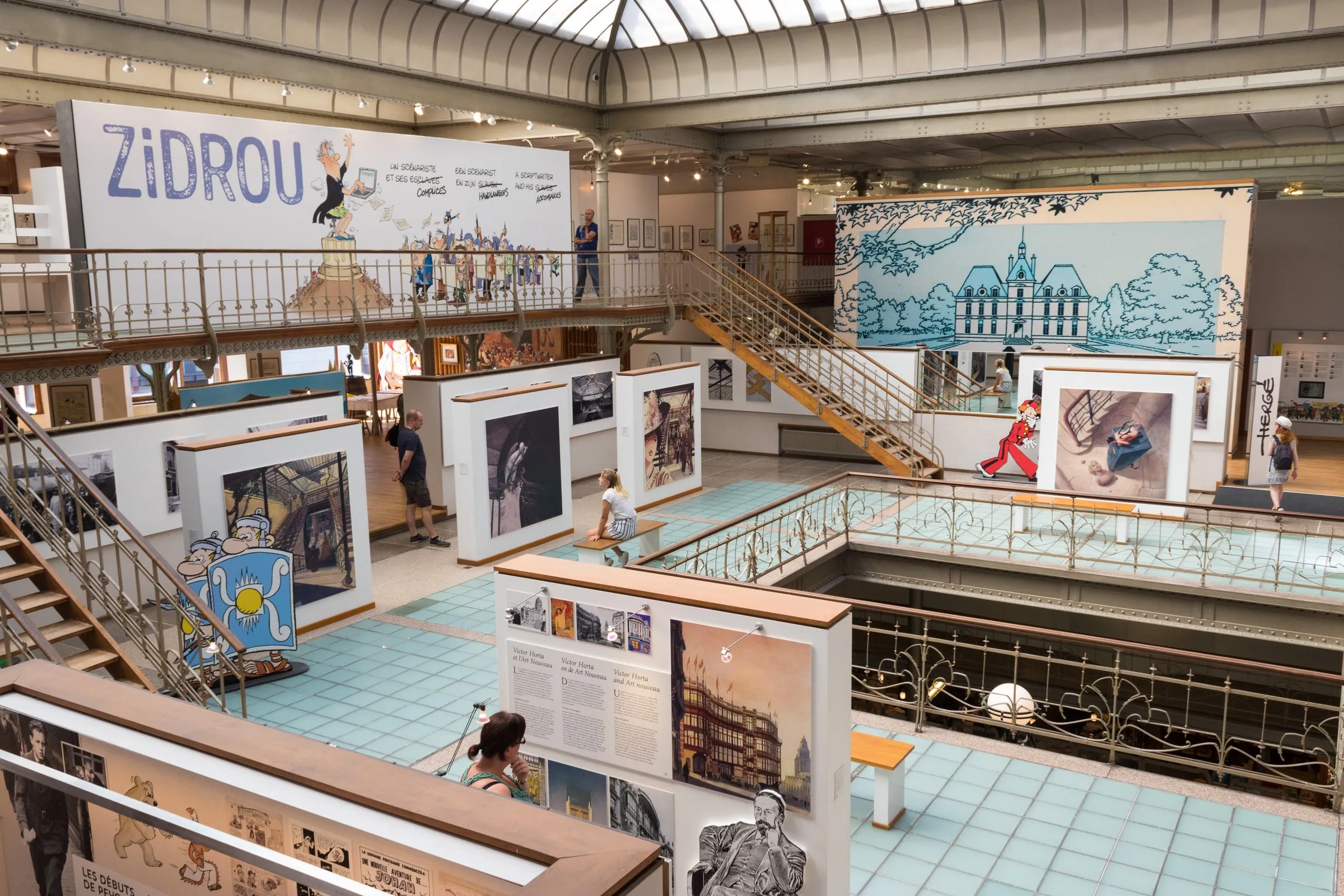 Inside the Belgian Comic Strip Center, a wrought-iron staircase and glass roof under Victor Horta's 1906 Art Nouveau ironwork, with exhibition panels below