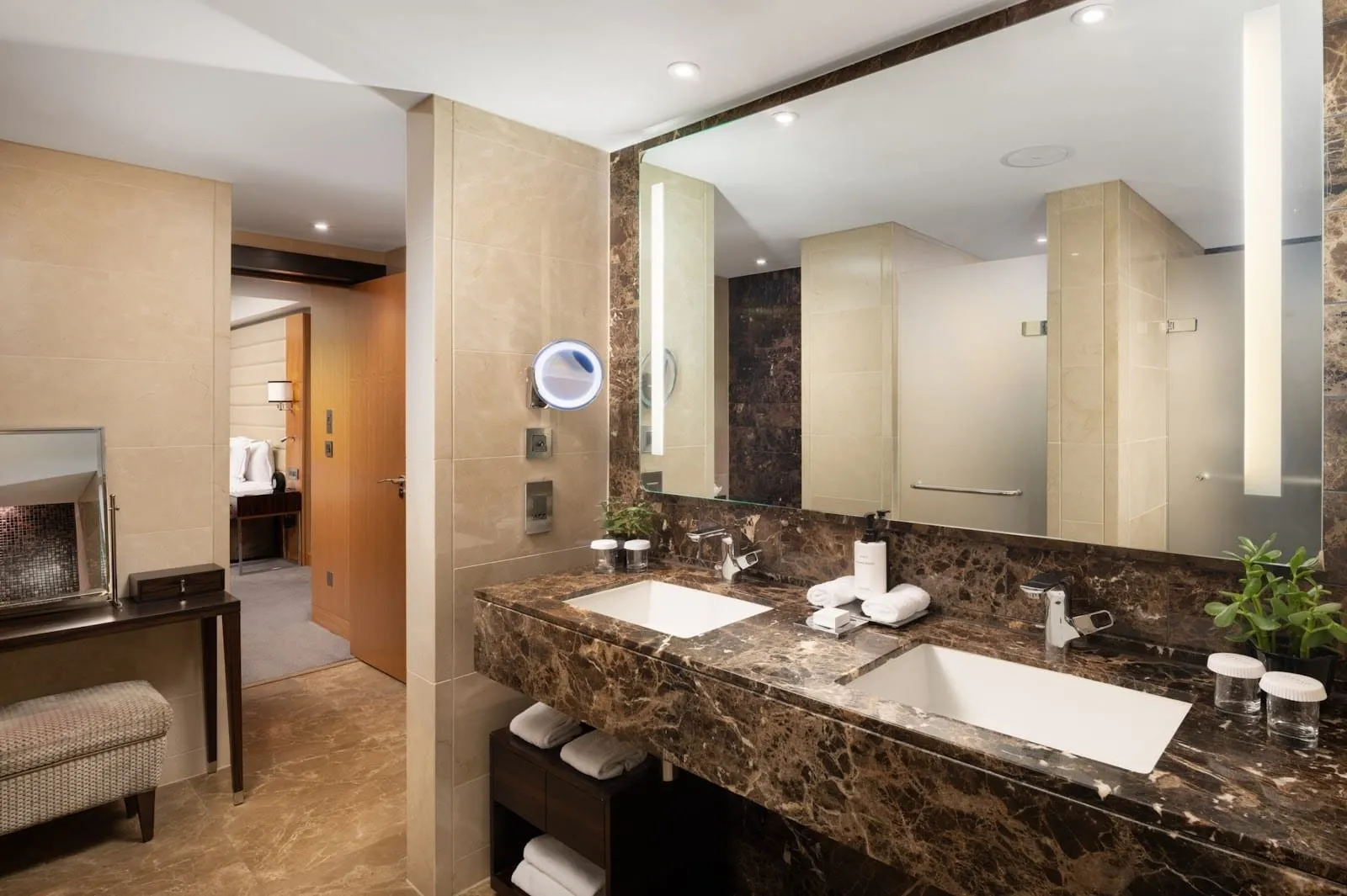 A bathroom at Conrad London St. James with twin basins set in a dark brown marble counter, a long lit mirror and cream stone walls, with the bedroom visible through the door