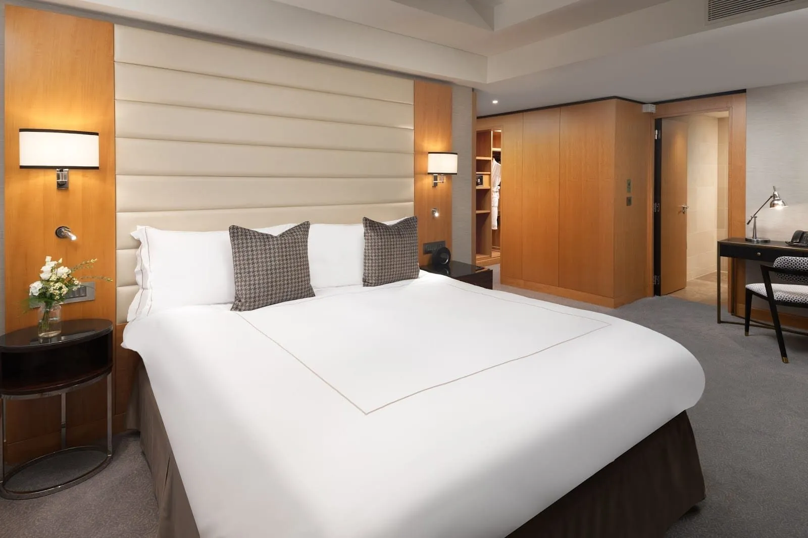 A room at Conrad London St. James with a cream quilted leather headboard between wood panels, houndstooth cushions, and an open walk-in wardrobe with robes beside the bathroom door