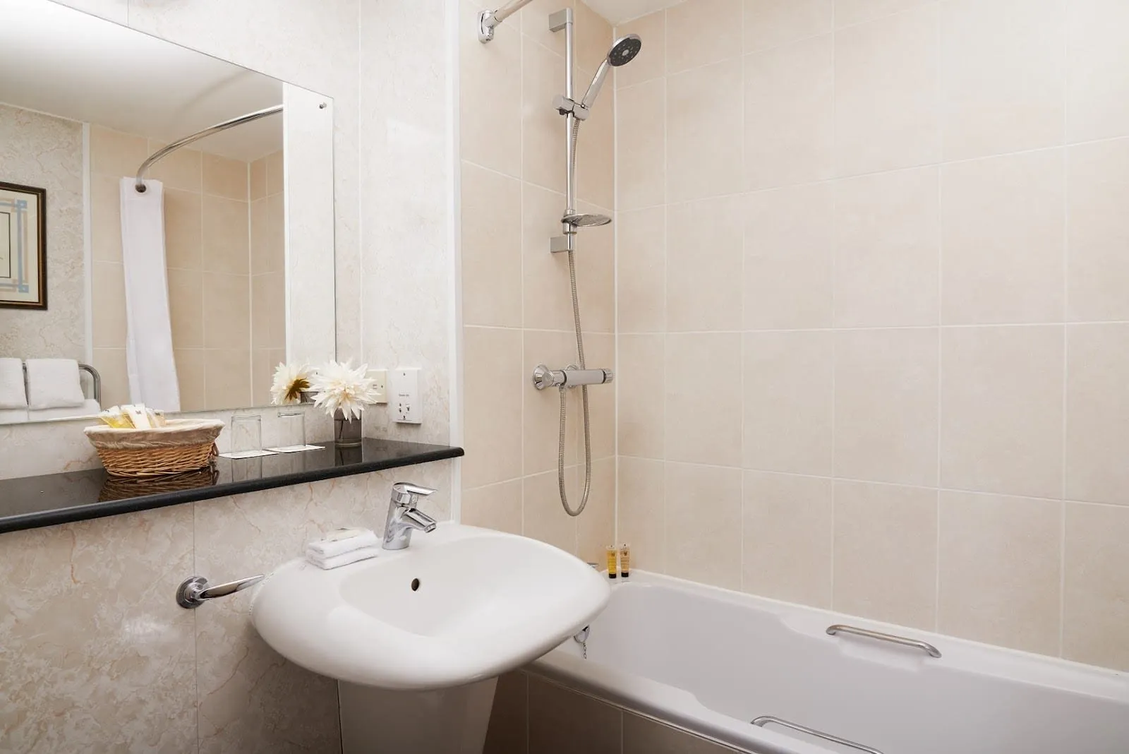 A cream-tiled bathroom at the Copthorne Tara with a pedestal basin, a wicker basket of toiletries and a bath with a grab rail and a shower over it