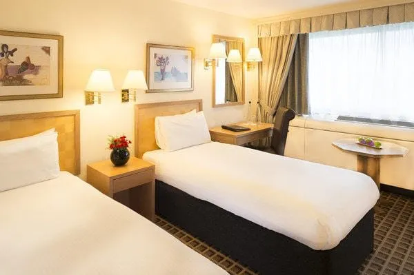 A twin room at the Copthorne Tara with two single beds against pale wood headboards, framed prints, brass wall lamps and a small table under a wide net-curtained window