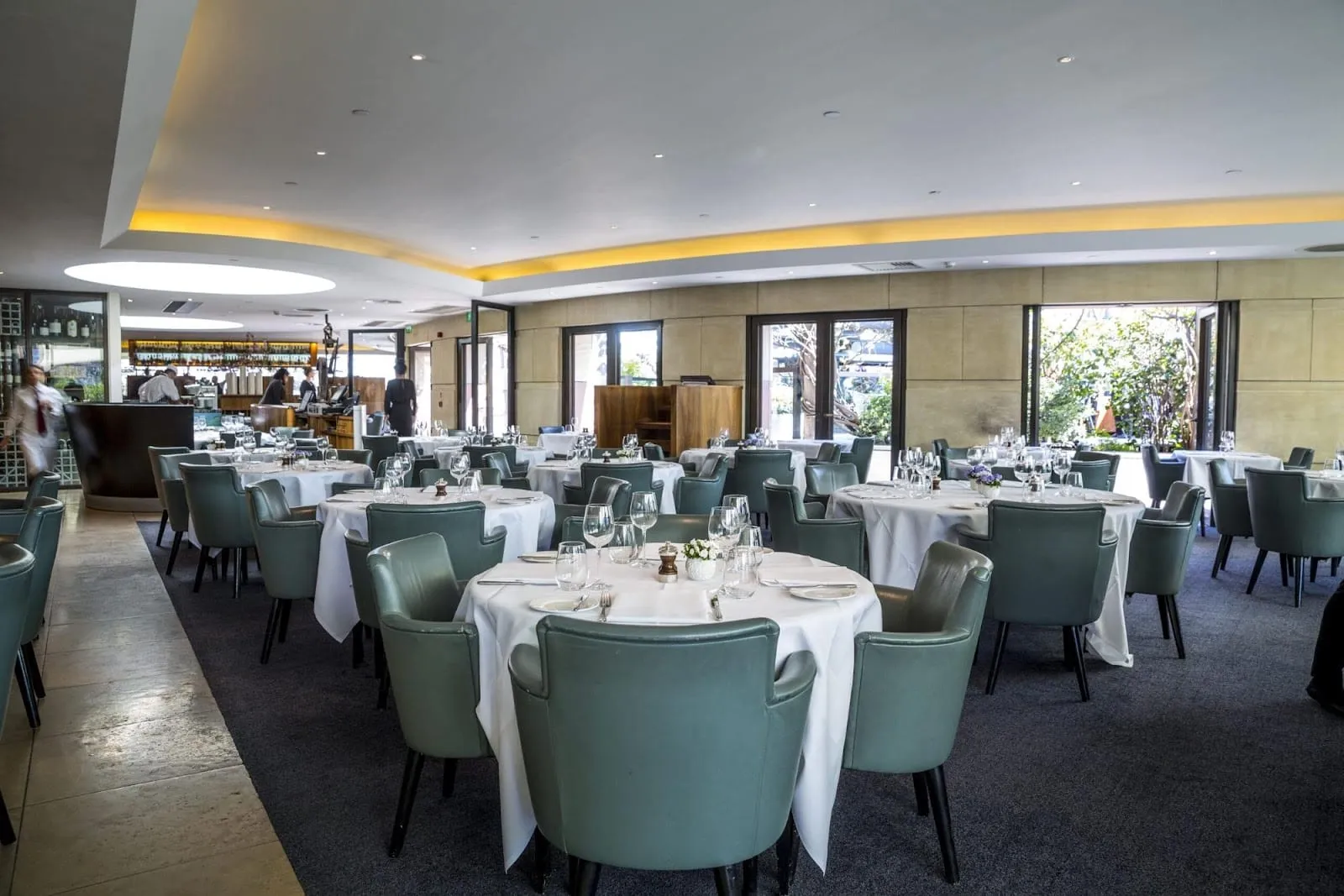 The indoor dining room at Coq d'Argent: round tables under white cloths with sage-green leather tub chairs, a bar along the far wall, and full-height glass doors standing open onto the planting of the roof garden