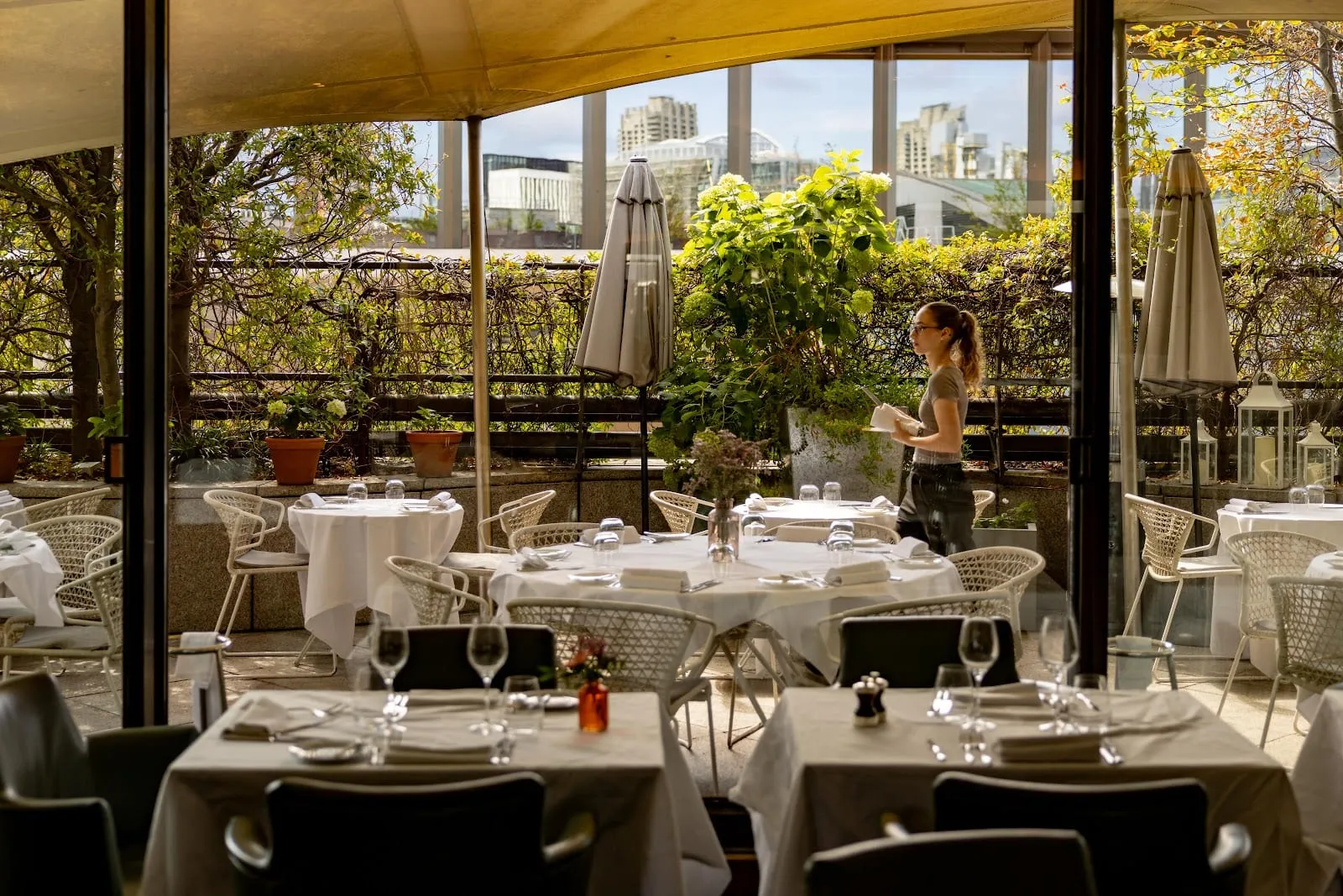 A rooftop restaurant terrace with white-clothed tables and rattan chairs under furled parasols, climbing greenery along the parapet and City buildings beyond