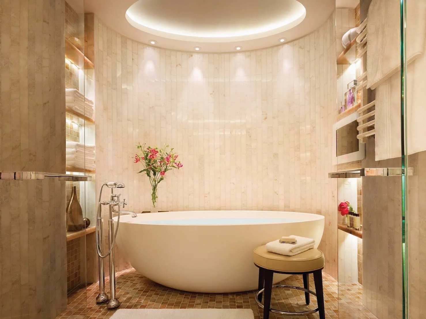 A suite bathroom at Corinthia London with a freestanding oval bath in a curved marble alcove under a lit ceiling dome, with a floor-standing tap and lit shelves of towels