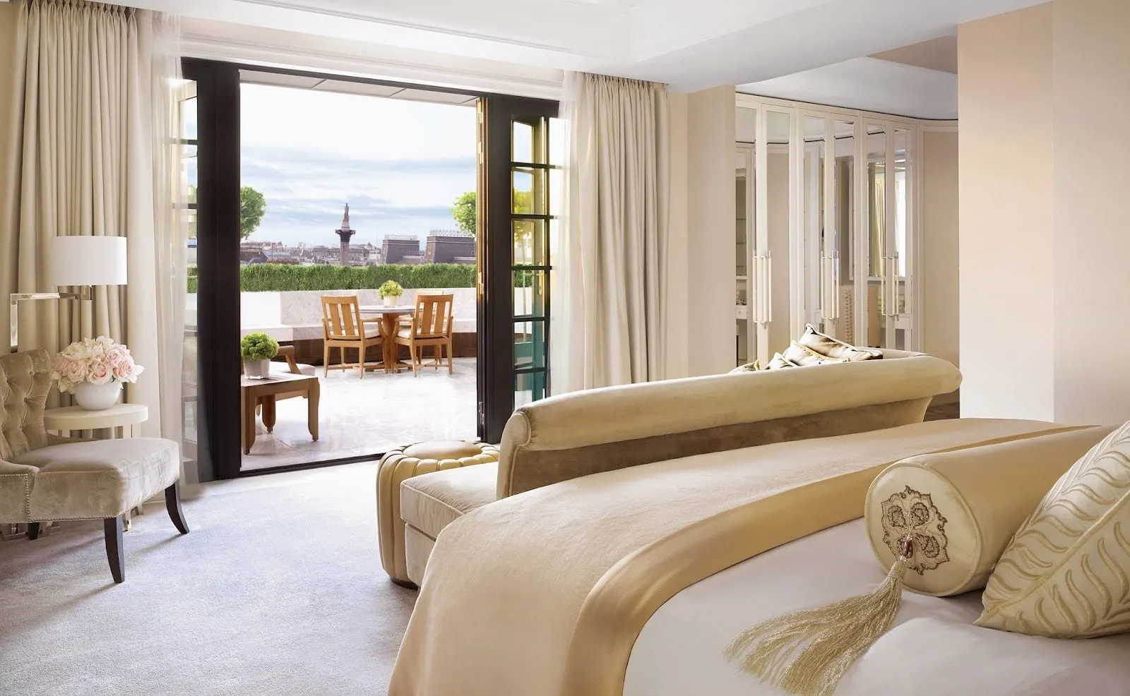 A suite bedroom at Corinthia London in cream and gold, with a curved velvet sofa at the foot of the bed and glass doors open onto a roof terrace with Nelson's Column beyond
