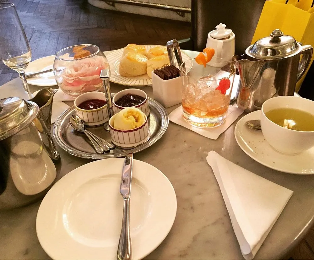 Warm scones with pots of jam, curd and clotted cream on a marble table, beside a silver teapot and a cup of green tea
