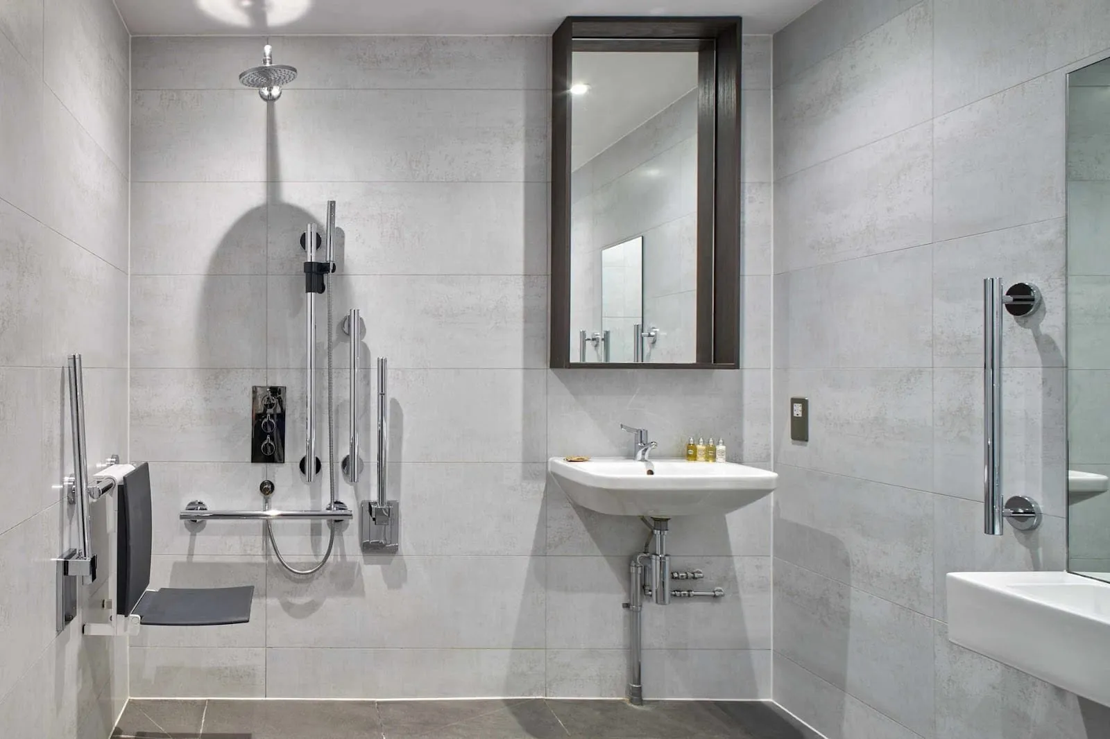 An accessible wet room at Cove Cannon Street in pale grey tiles, with a fold-down shower seat, grab rails, a rain shower and a wall-hung basin