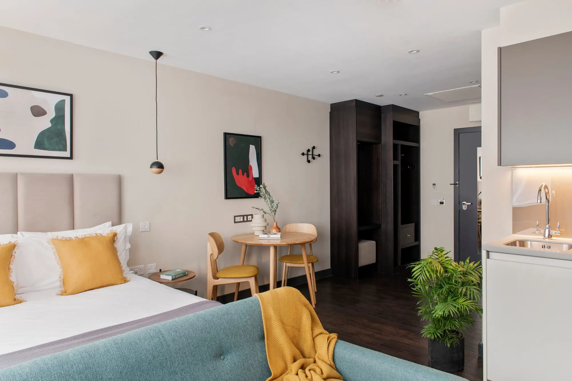 A studio at Cove Cannon Street, with a mustard-cushioned bed, a two-seat dining table and a kitchenette sink against the far wall