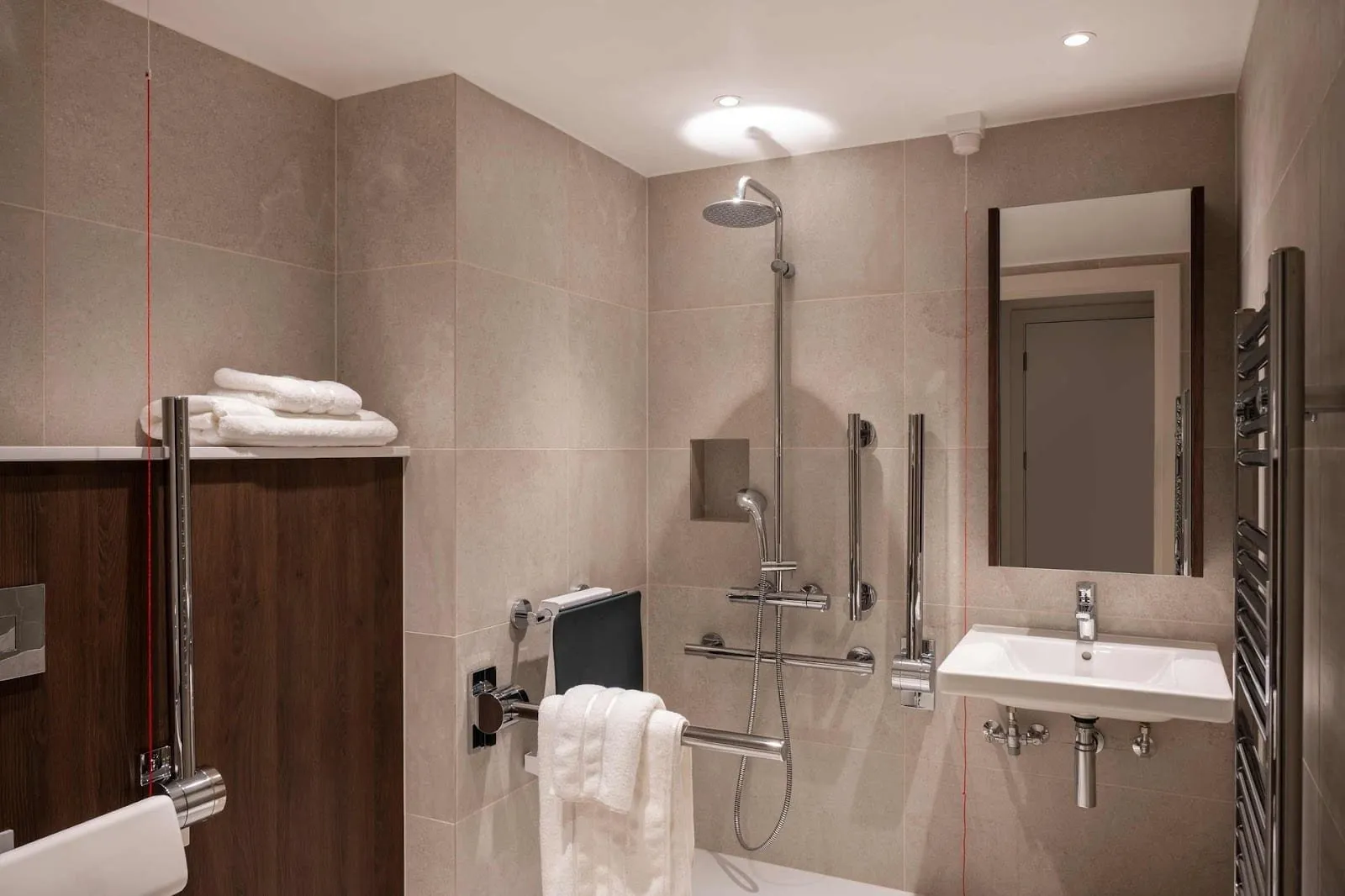 An accessible wet room at Cove Landmark Pinnacle with a walk-in rainfall shower, a fold-down shower seat, chrome grab rails, a red emergency pull cord and a wall-mounted basin