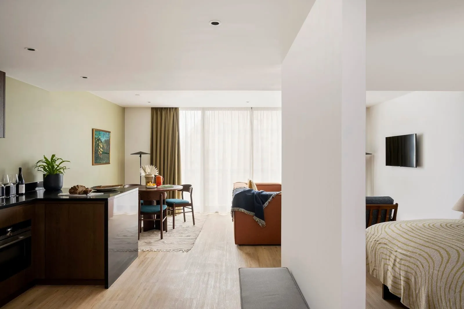 An open-plan apartment at Cove Landmark Pinnacle: a dark wood kitchen with a built-in oven and black worktop, a round dining table with teal chairs, a rust-coloured sofa by full-height curtained windows, and a bed behind a partition wall