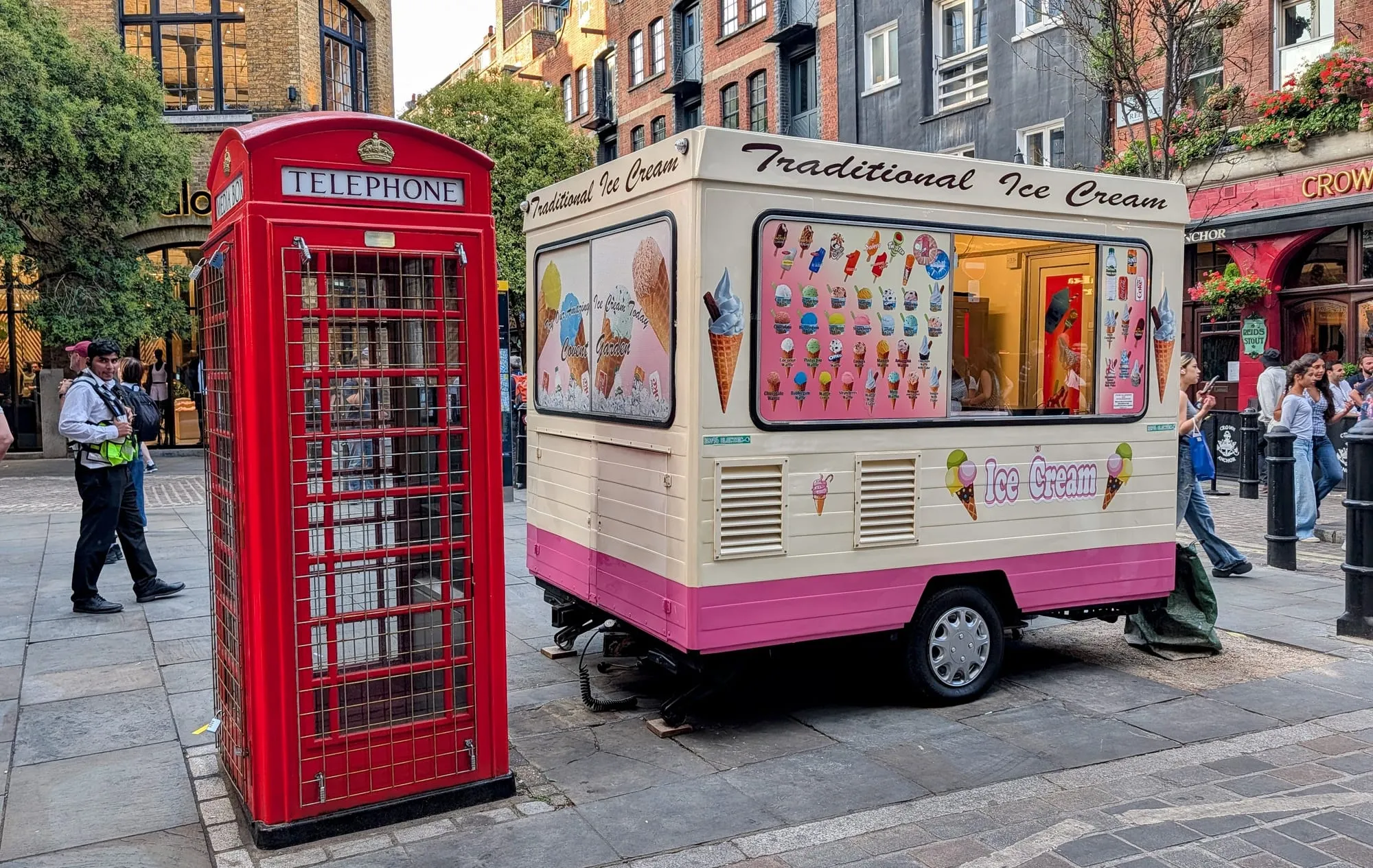 A traditional ice cream van painted cream and pink parked beside a red telephone box on the cobbles at Covent Garden, its menu board covered in cones and lollies