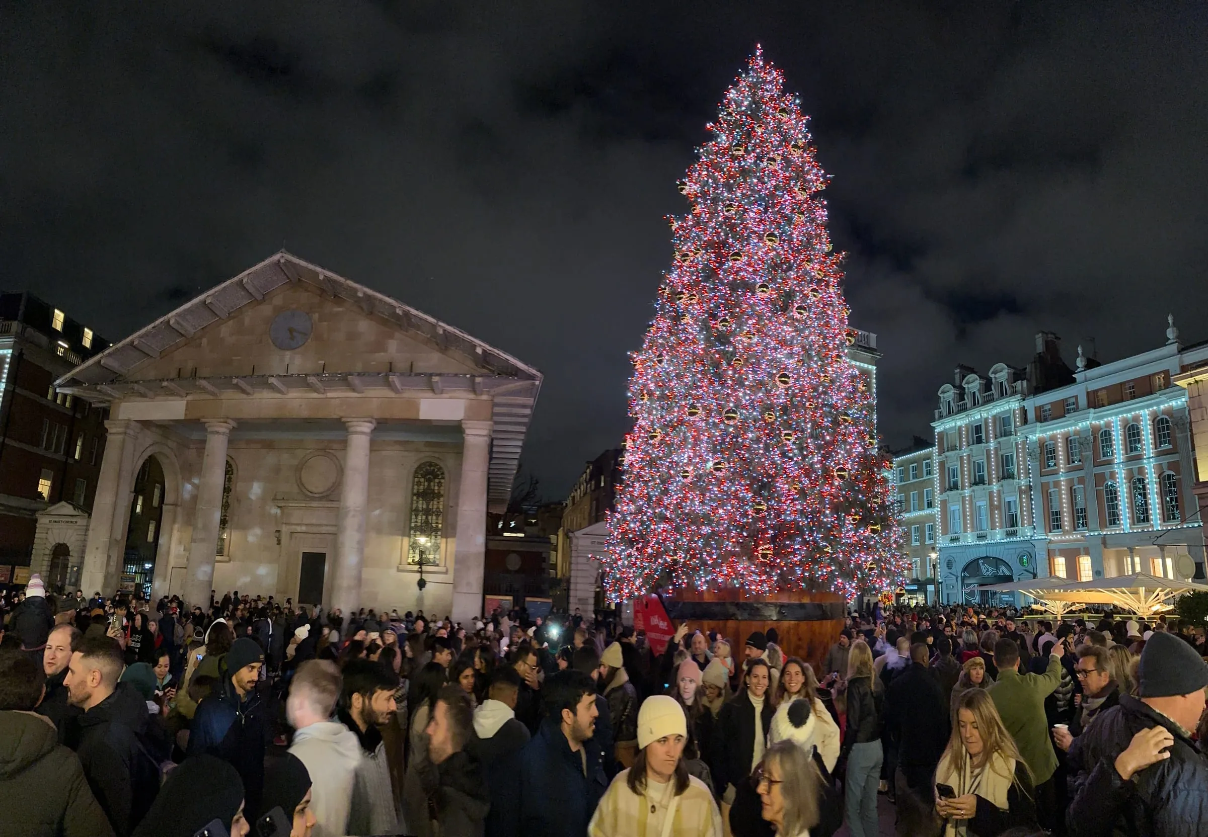 A tall Christmas tree covered in red and white lights on Covent Garden's West Piazza beside the portico of St Paul's Church, with a large crowd in front