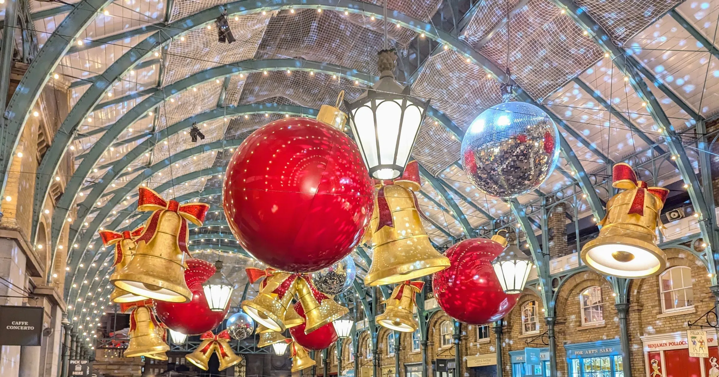 Giant gold bells tied with red bows, red baubles and a mirror ball hanging under the lit iron roof of Covent Garden's Market Building, with the shopfronts of the market hall behind