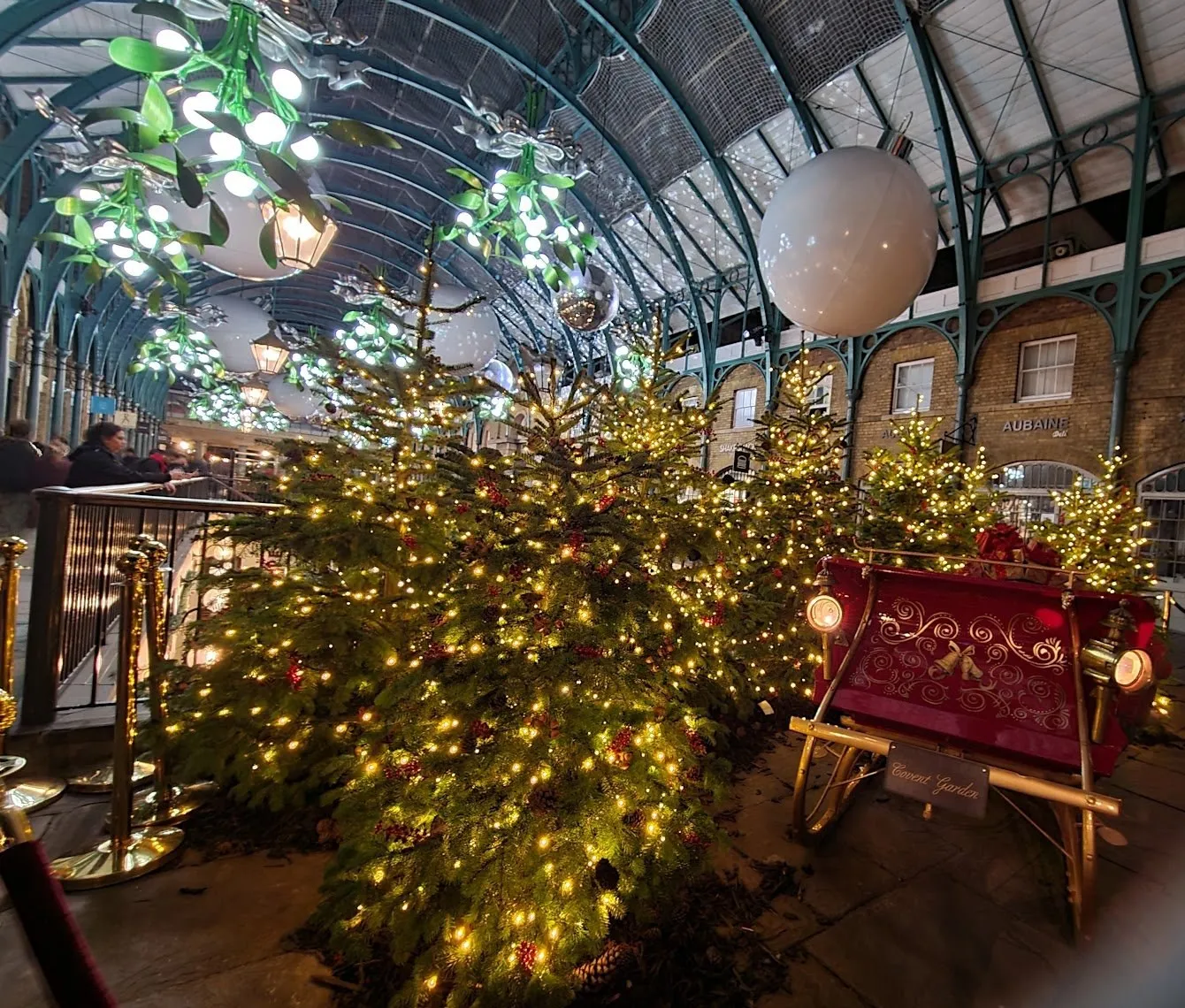 Inside Covent Garden's Market Building, lit Christmas trees and a red sleigh under the iron roof, with giant white baubles, mirror balls and light-up mistletoe hanging above