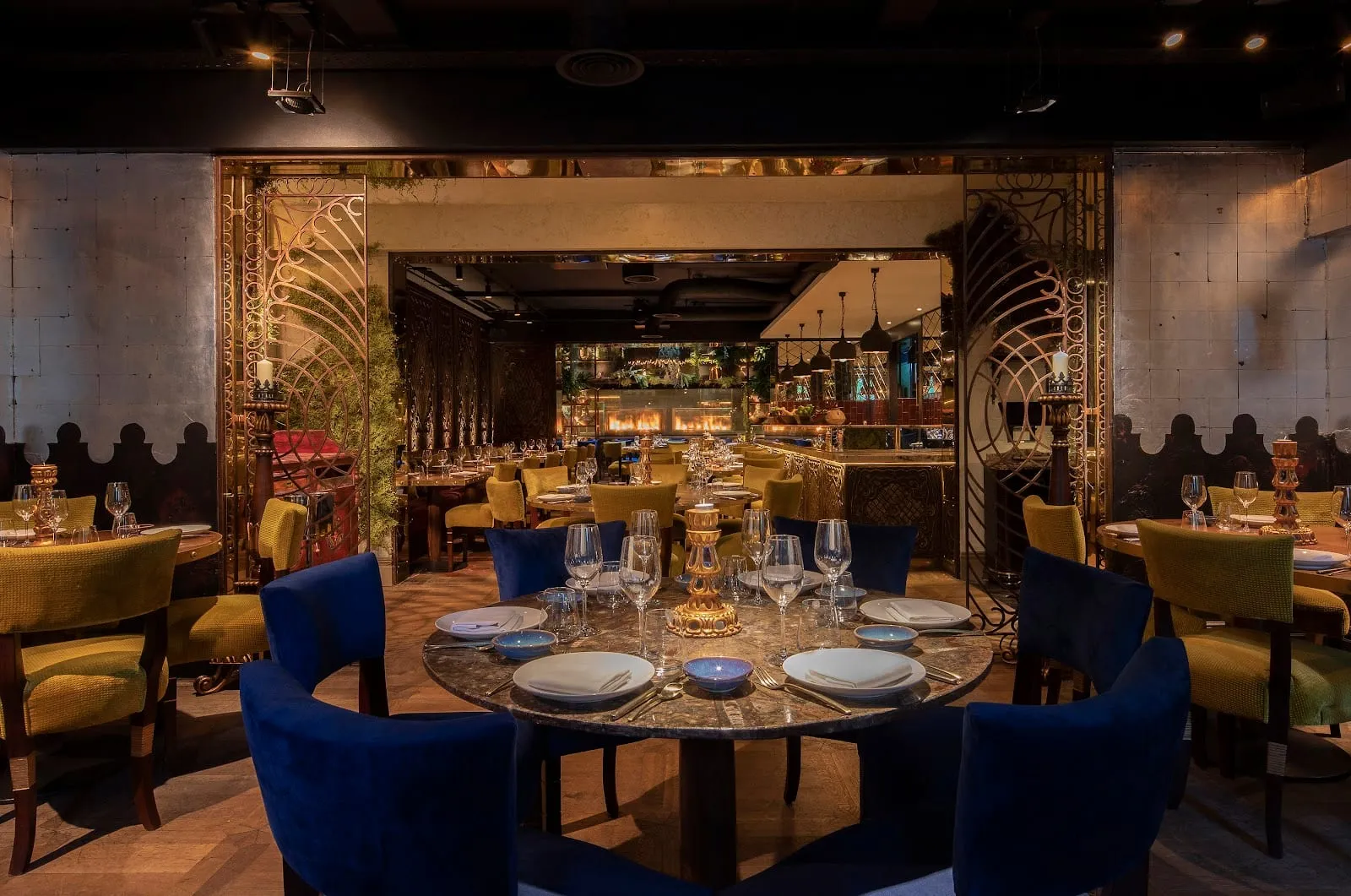 A low-lit dining room with gilded ironwork screens, olive-green and blue velvet chairs, a round marble table laid for six, and flames in an open kitchen beyond
