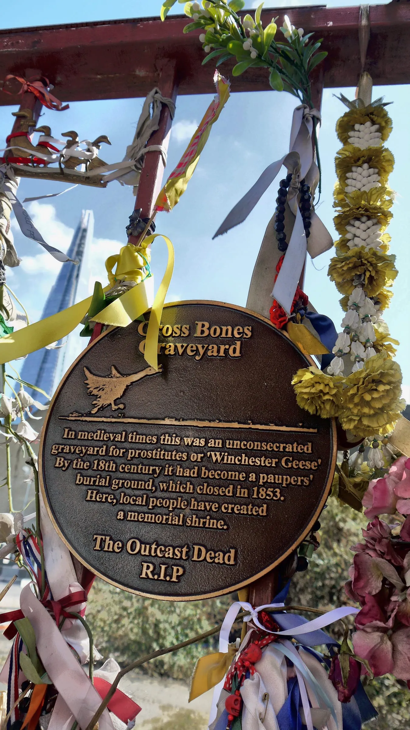 A bronze plaque reading Cross Bones Graveyard, tied round with ribbons, flowers and garlands on the red iron gates, with the Shard behind