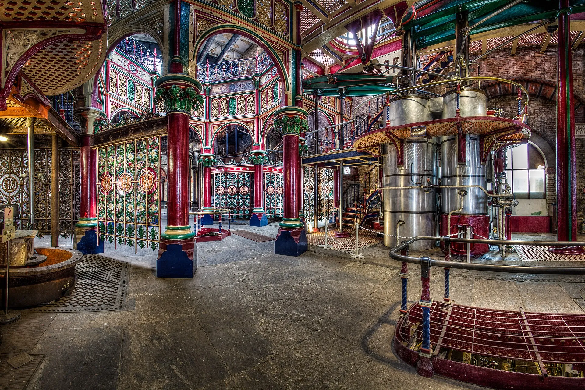 The Beam Engine House at Crossness Pumping Station, its cast ironwork painted in red, green and gold around an octagonal screen, with the polished cylinders of the Prince Consort engine and its nameplate on the right