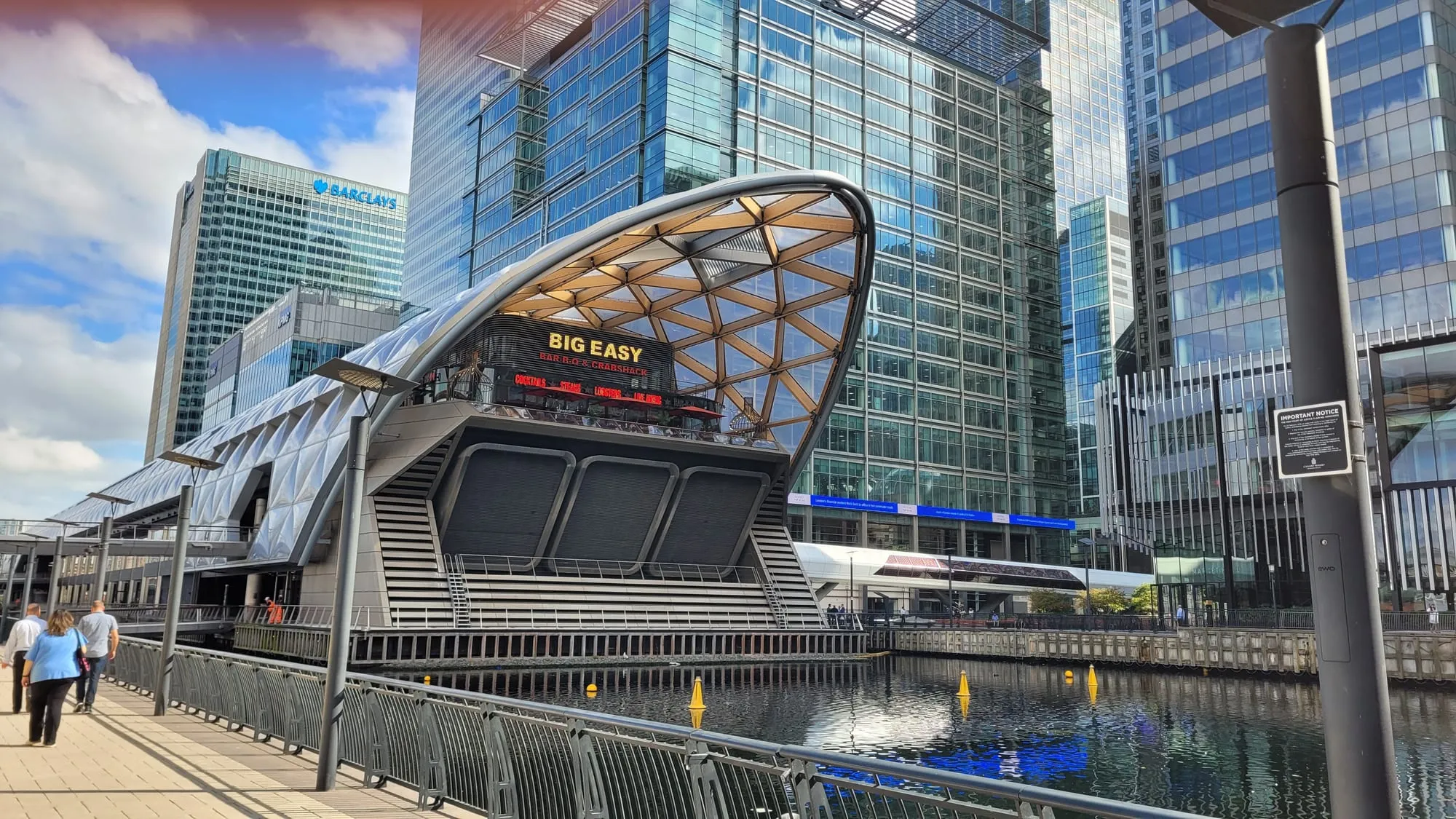 The timber lattice roof of Crossrail Place rising out of the dock water at Canary Wharf, with office towers behind