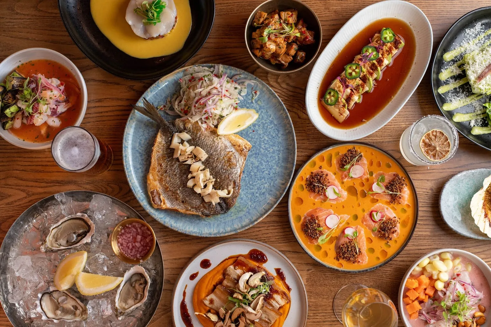 An overhead spread of ceviches in cloudy and orange marinades, a whole grilled bream under shaved onion salad, oysters on ice, scallops in yellow sauce and pork belly on orange purée, on a wooden table