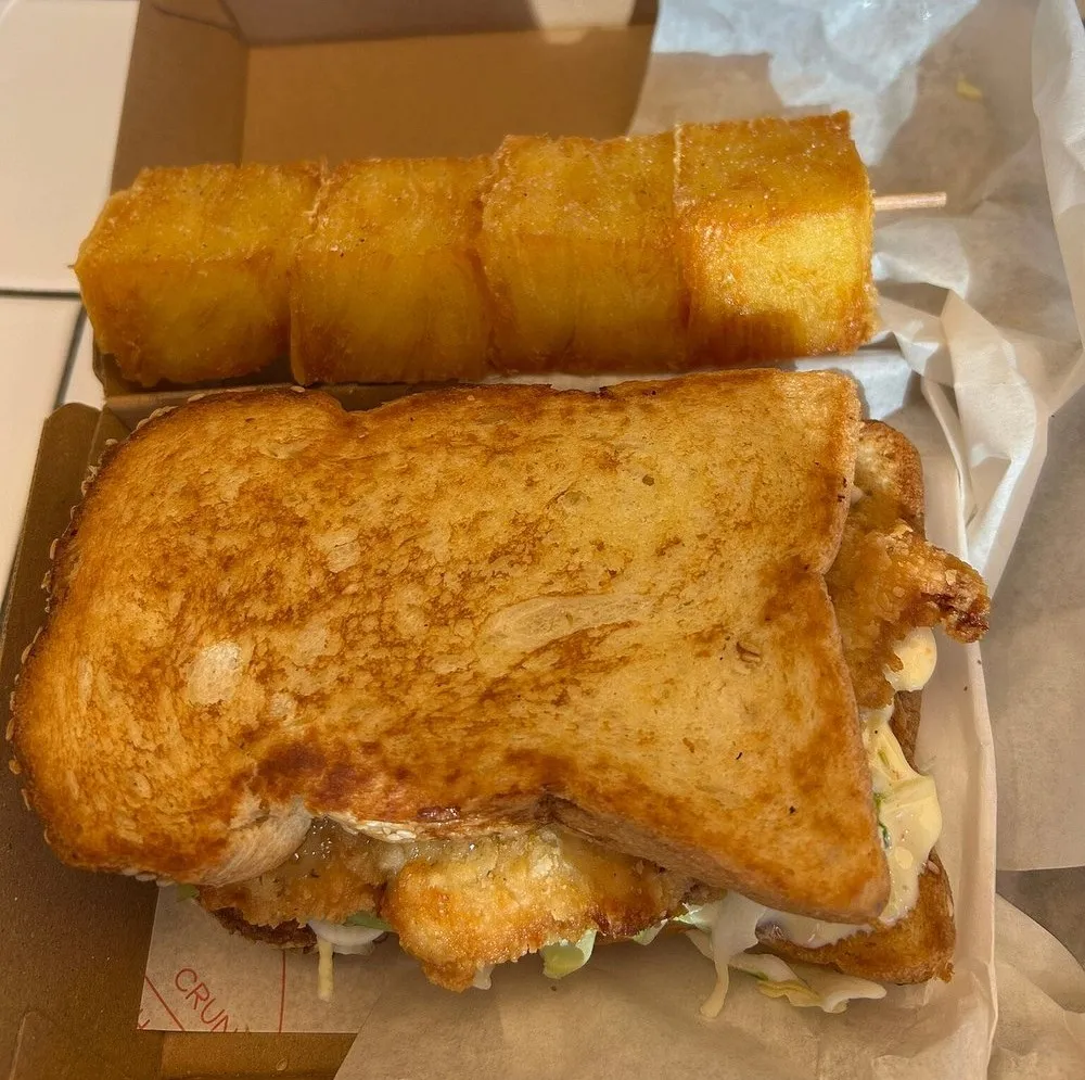 A fried chicken sandwich in thick golden brioche, in a takeaway box beside a skewer of fried potato