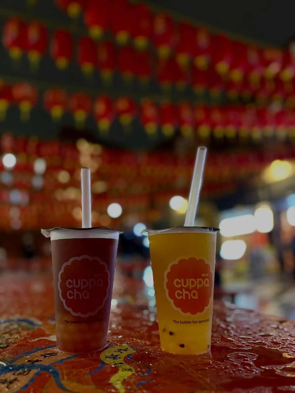 Two Cuppacha cups, one deep red and one orange, standing on a table under a canopy of red lanterns