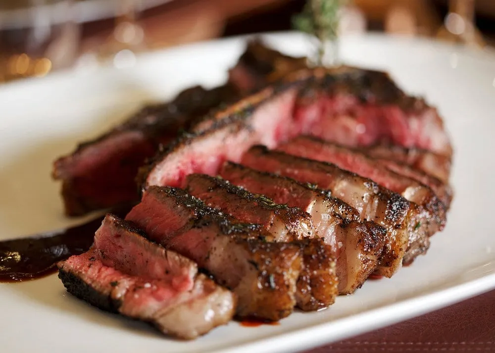 A sliced steak at CUT, dark and heavily crusted at the edge with a red centre, fanned across a white plate with a sprig of thyme and a dark sauce