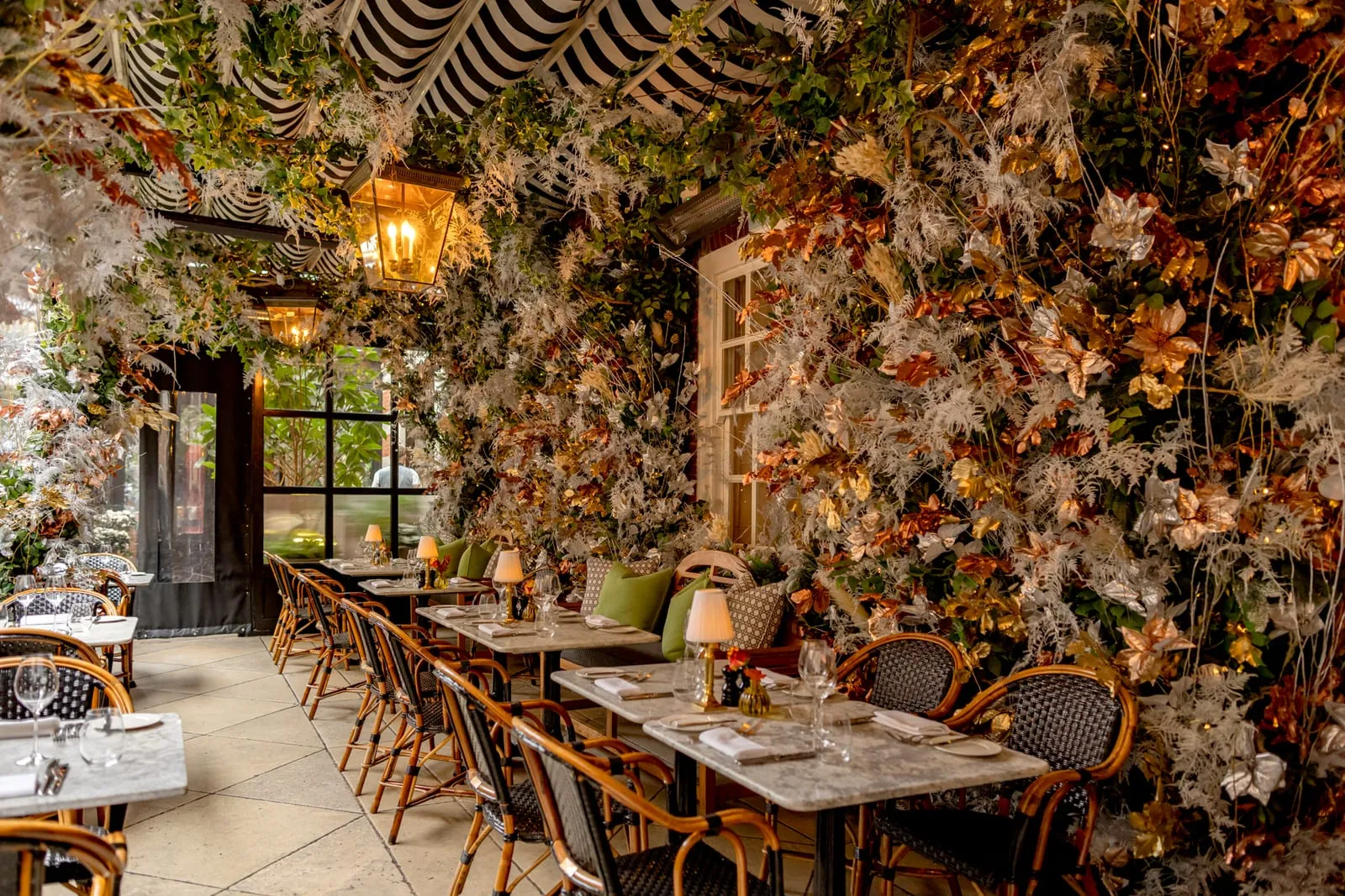 Dalloway Terrace dressed in dried autumn foliage in copper, cream and gold across the walls and ceiling, with marble tables and rattan chairs